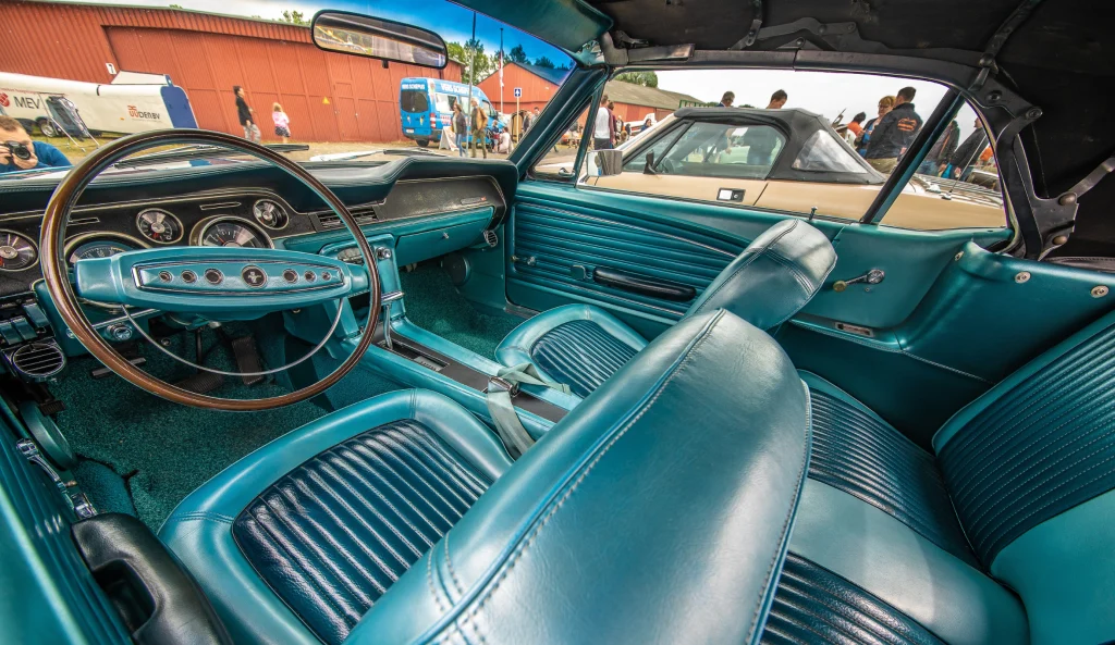 Visão interna detalhada do cockpit de um carro clássico conversível com acabamento e bancos de couro na cor azul-turquesa. Em destaque, o volante antigo com detalhes cromados, o painel de instrumentos analógicos e os bancos dianteiros e traseiros. Pela janela, vê-se um evento de carros ao ar livre com pessoas e galpões ao fundo.